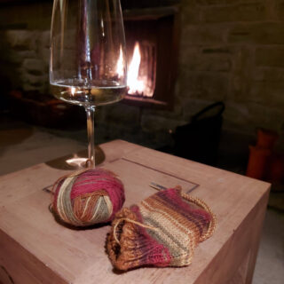 A glass of wine and partly-knitted sock in shades of red, green and brown sit on a wooden coffee table. In the background is an open fire.