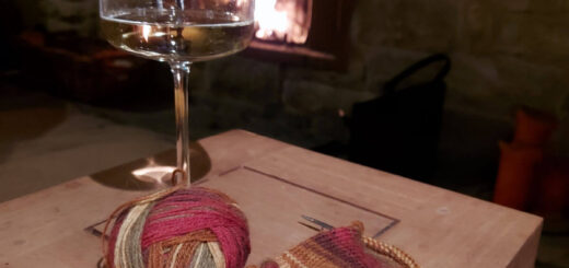 A glass of wine and partly-knitted sock in shades of red, green and brown sit on a wooden coffee table. In the background is an open fire.