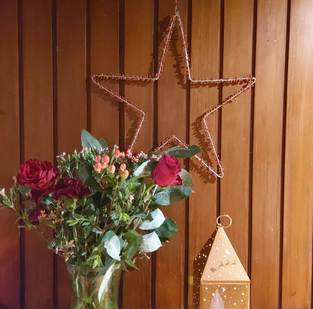 A large star decorated with berries hangs on a wood-panelled wall. Below is a vase of flowers and a bronze-coloured Christmas decoration