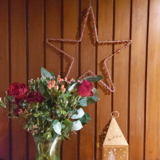 A large star decorated with berries hangs on a wood-panelled wall. Below is a vase of flowers and a bronze-coloured Christmas decoration