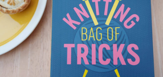A book called Patty Lyons' Knitting Bag of Tricks lies on a wooden coffee table next to an orange mug of tea and a mince pie on a yellow plate