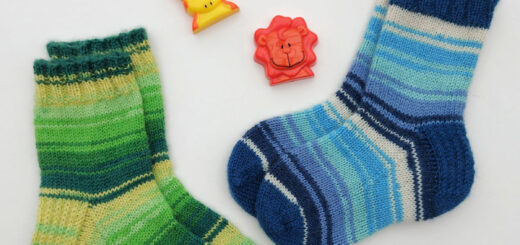 Two pairs of hand-knitted children's socks against a white background. The pair on the left is slightly bigger and is shades of green stripes; the pair on the right is in shades of blue stripes with blue cuffs, heels and toes. There are three toy shapes lying next to the socks - two lions and a giraffe