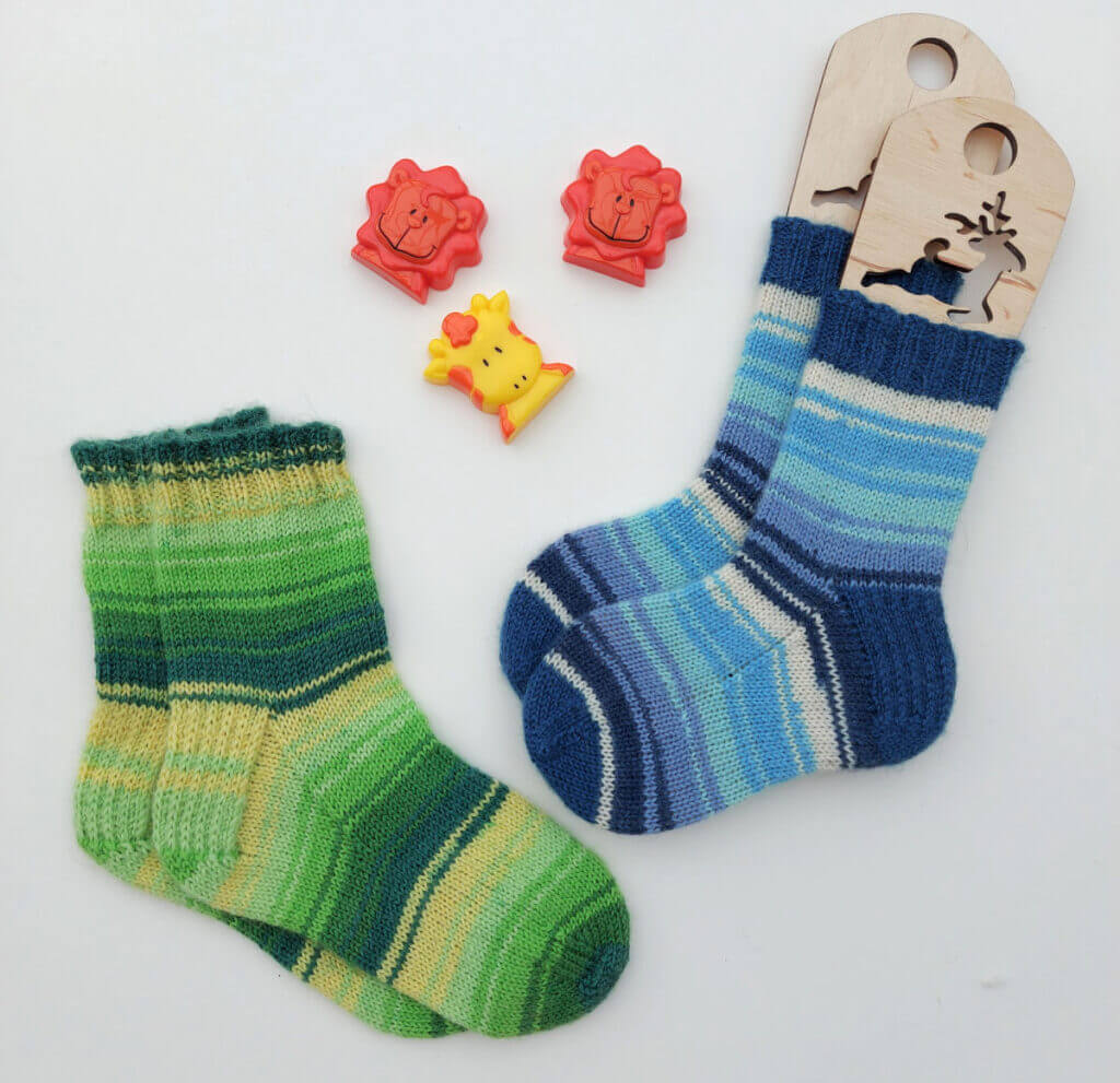 Two pairs of hand-knitted children's socks against a white background. The pair on the left is slightly bigger and is shades of green stripes; the pair on the right is in shades of blue stripes with blue cuffs, heels and toes. The blue pair is on a set of wooden sock blockers. There are three toy shapes lying next to the socks - two lions and a giraffe