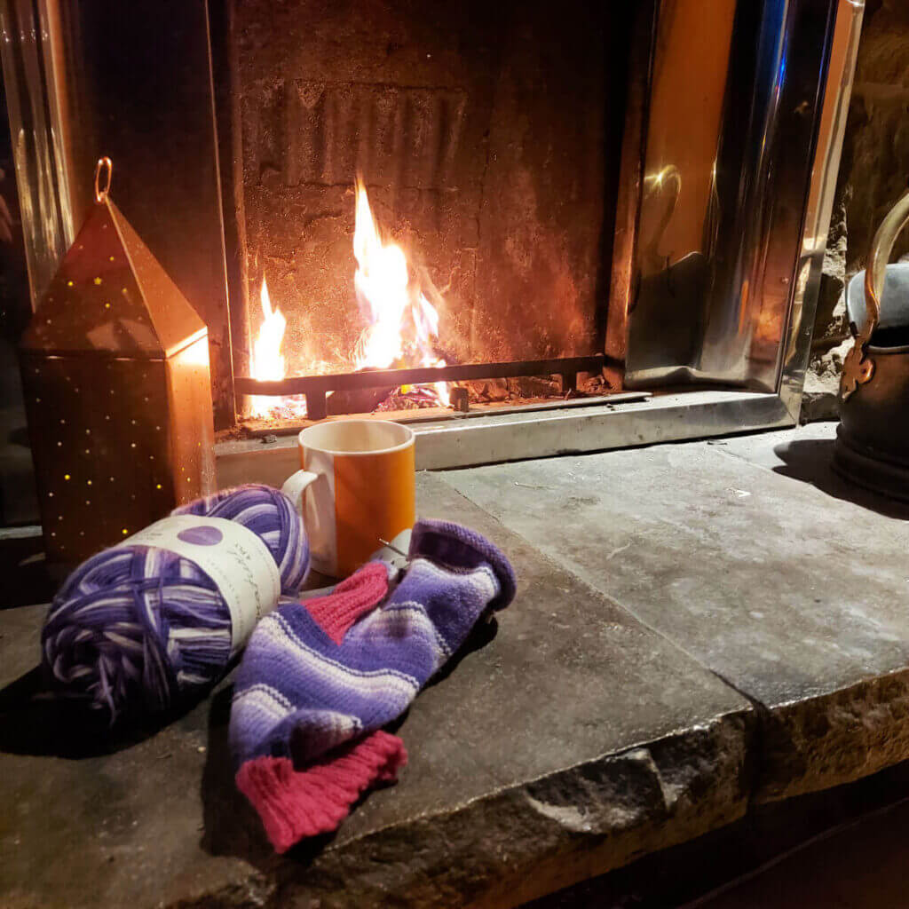 A partly-knitted purple striped sock with a pink cuff and heel lies on a stone hearth in front of an open fire. Next to it is a bronze light decoration.
