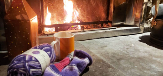 A partly-knitted purple striped sock with a pink cuff and heel lies on a stone hearth in front of an open fire. Next to it is a bronze light decoration.