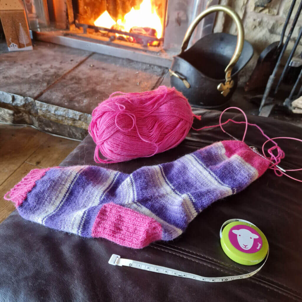 A purple striped sock with pink cuffs, heel and toes lies on a leather foot stool in front of an open fire