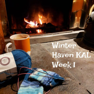 A partly-knitted sock and an orange mug in front of an open fire. Words on the photo read "Winter Haven KAL Week 1"