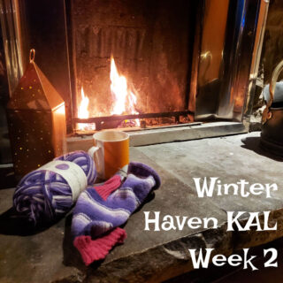 A partly-knitted purple striped sock with a pink cuff and heel lies on a stone hearth in front of an open fire. Next to it is a bronze light decoration. The words "Winter Haven KAL Week 2" are at the bottom right of the photo