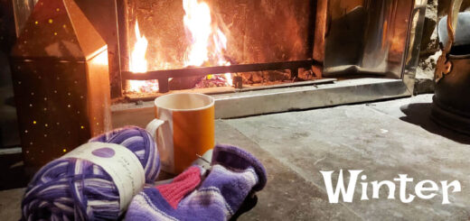 A partly-knitted purple striped sock with a pink cuff and heel lies on a stone hearth in front of an open fire. Next to it is a bronze light decoration. The words "Winter Haven KAL Week 2" are at the bottom right of the photo