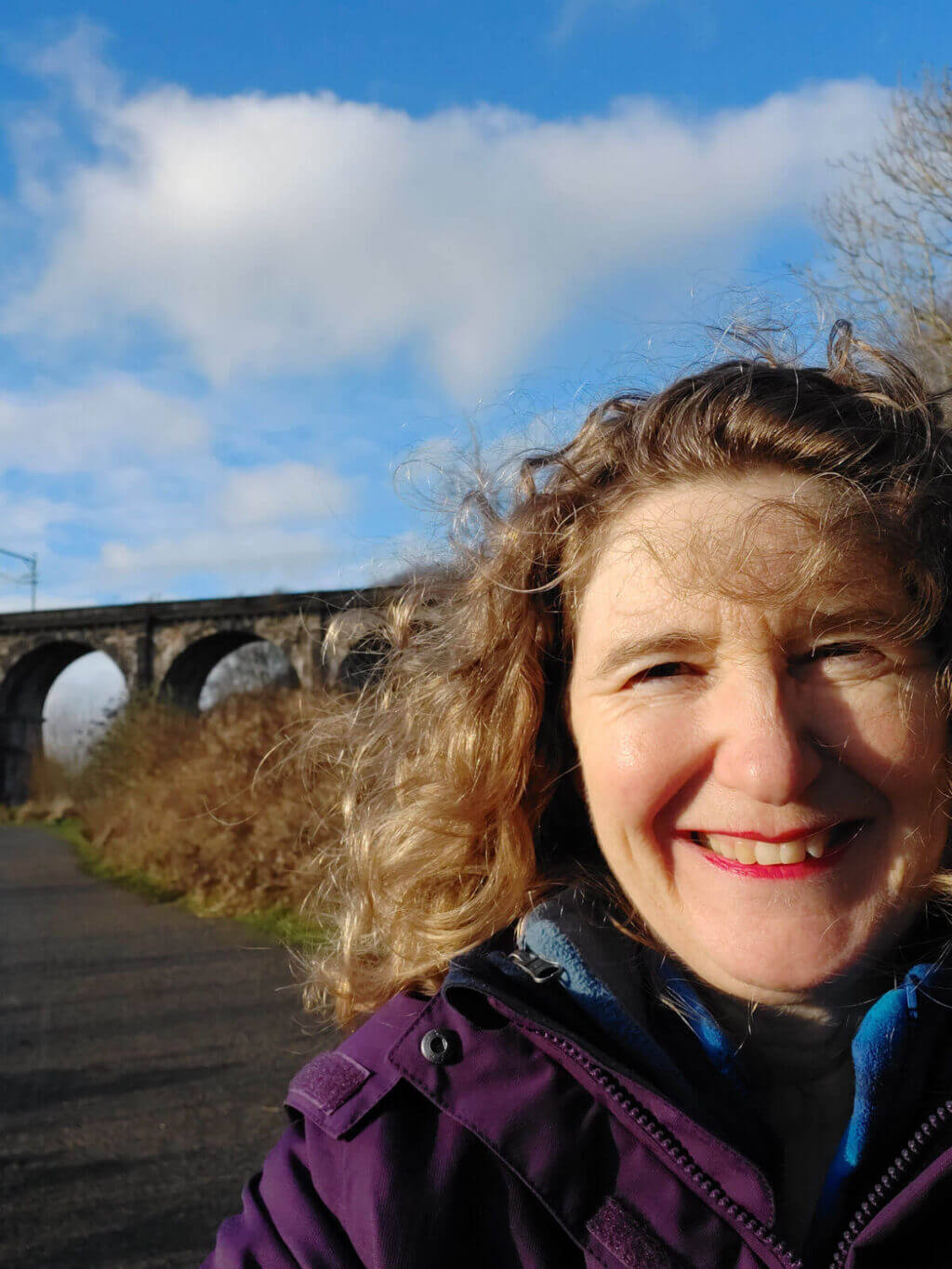 A woman with blonde curly hair smiles at the camera. She is wearing a purple coat with a blue fleece lining and behind her is a blue sky and brick railway arches