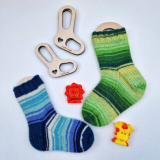 A flatlay of two small socks on wooden blockers; one sock is blue stripes and the other is green stripes. Above them on the white background are two more tiny blockers without socks on them, and between the socks are an orange lion toy and a yellow giraffe toy