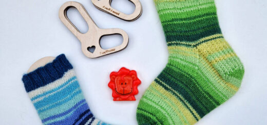 A flatlay of two small socks on wooden blockers; one sock is blue stripes and the other is green stripes. Above them on the white background are two more tiny blockers without socks on them, and between the socks are an orange lion toy and a yellow giraffe toy