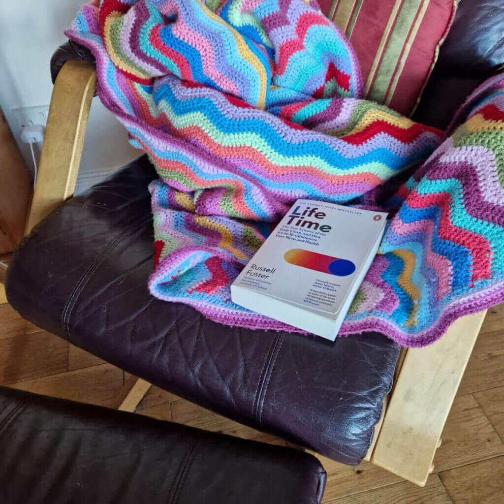 A brightly coloured crochet ripple blanket on a brown leather rocking chair. There is a book resting on the blanket