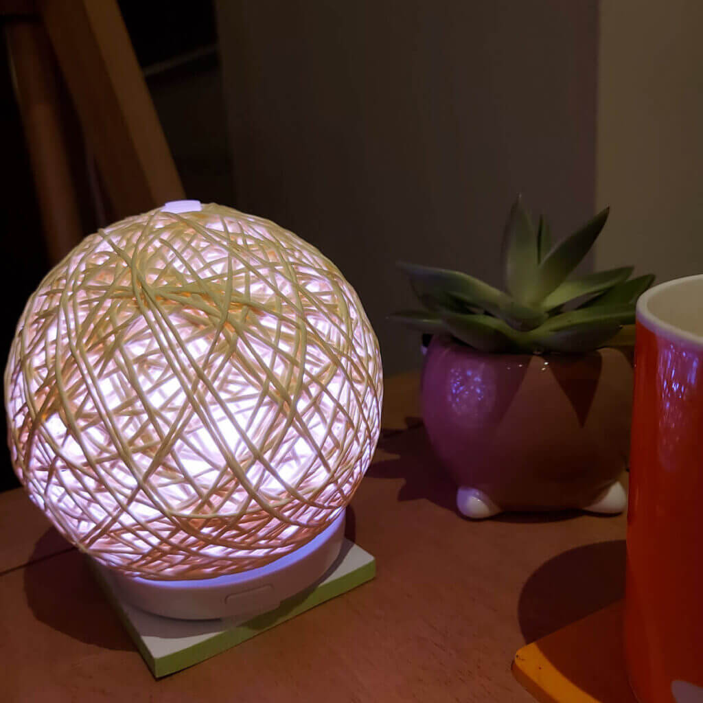 A round diffuser that looks like ball of yarn. It's glowing pink and sitting on a table next to an orange mug of tea and a succulent plant