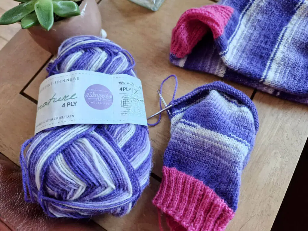 A partly-knitted purple striped sock with a pink cuff sits on a wooden table next to a ball of purple striped yarn and a completed sock in the same colours. A small plant in a ceramic pot sits behind the knitting