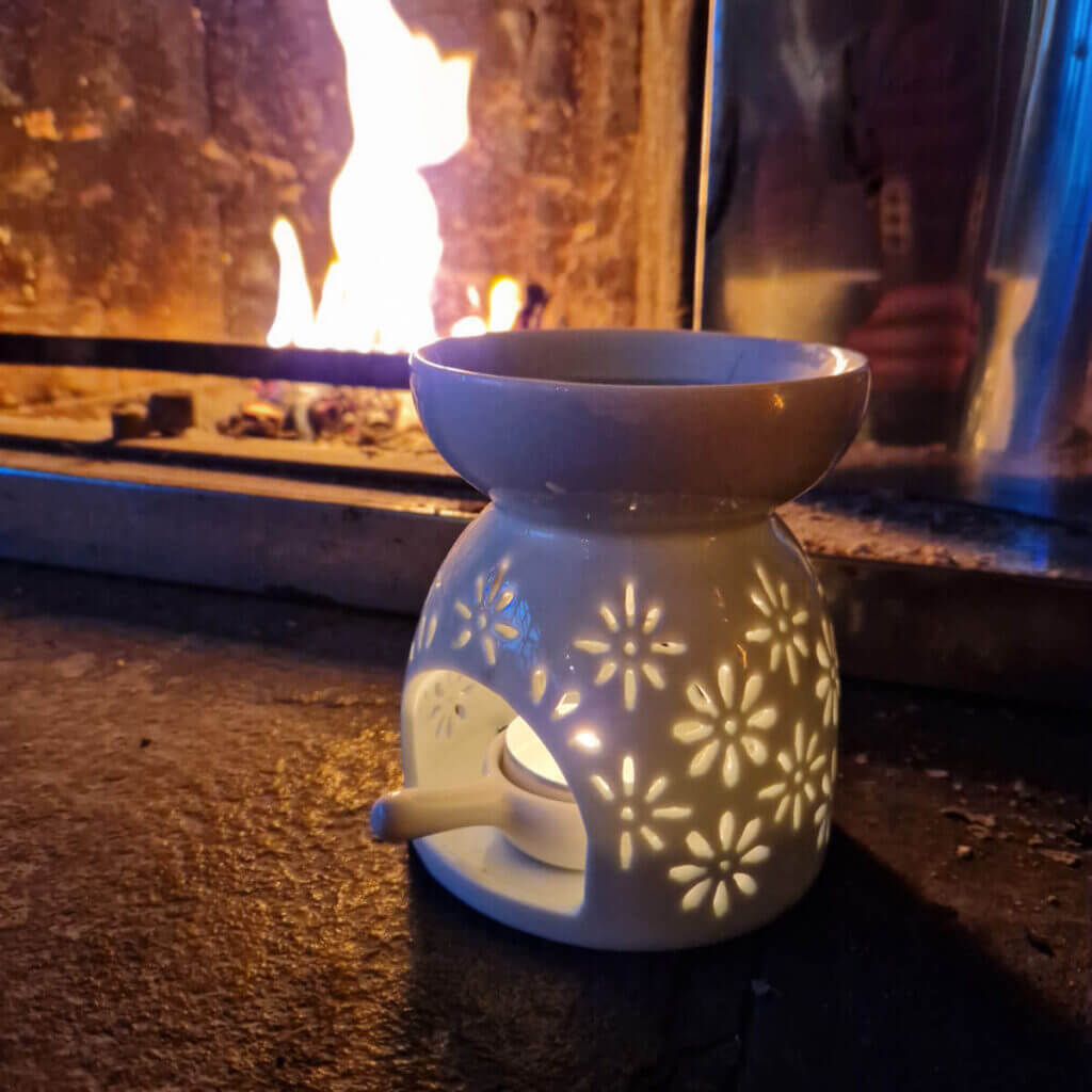 A wax melt burner with flower-shaped motifs around the side stands on a stone hearth in front of an open fire
