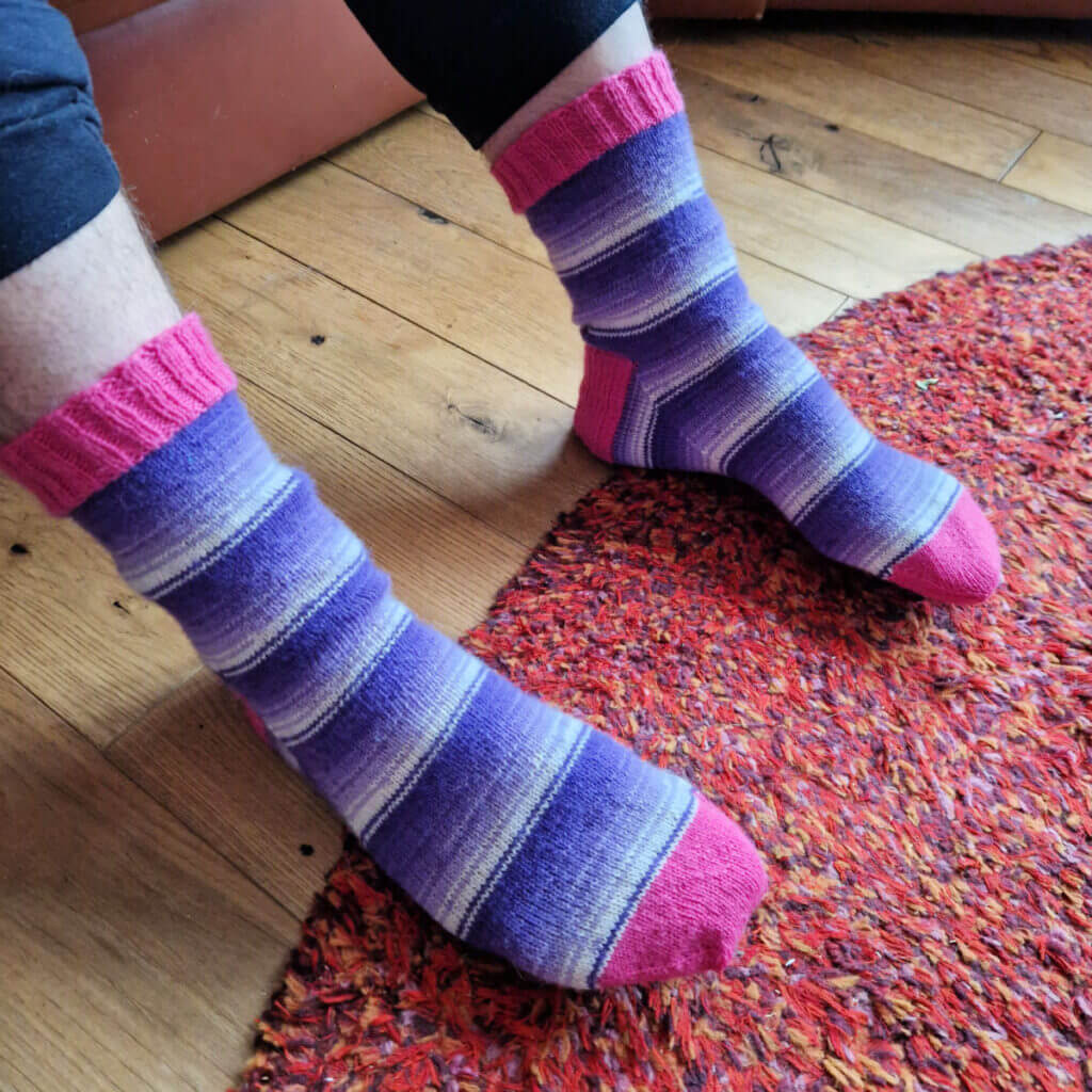 A pair of socks in purple and white stripes with pink cuffs, heels and toes are on a pair of feet.  There's a red rug on the wooden floor and the person wearing the socks is sitting on a brown leather sofa