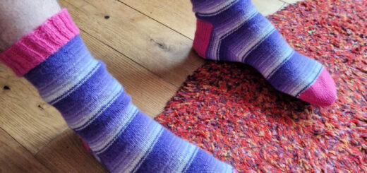 A pair of socks in purple and white stripes with pink cuffs, heels and toes are on a pair of feet. There's a red rug on the wooden floor and the person wearing the socks is sitting on a brown leather sofa