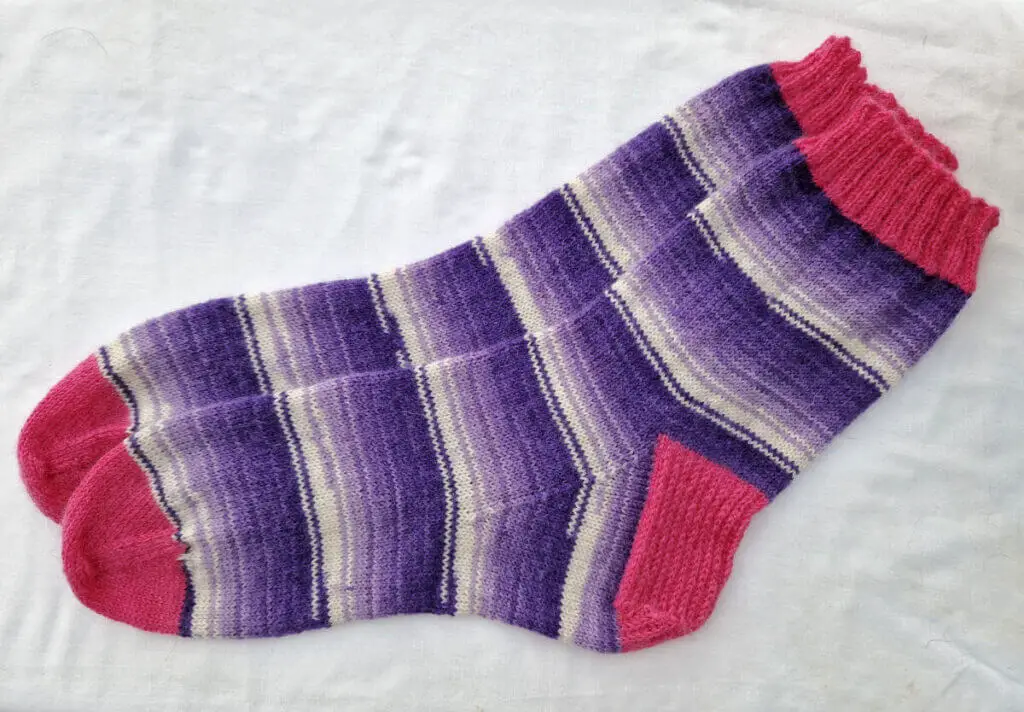 A pair of purple and white socks with pink cuffs, heels and toes lying on a white background. The socks have been blocked and look beautiful