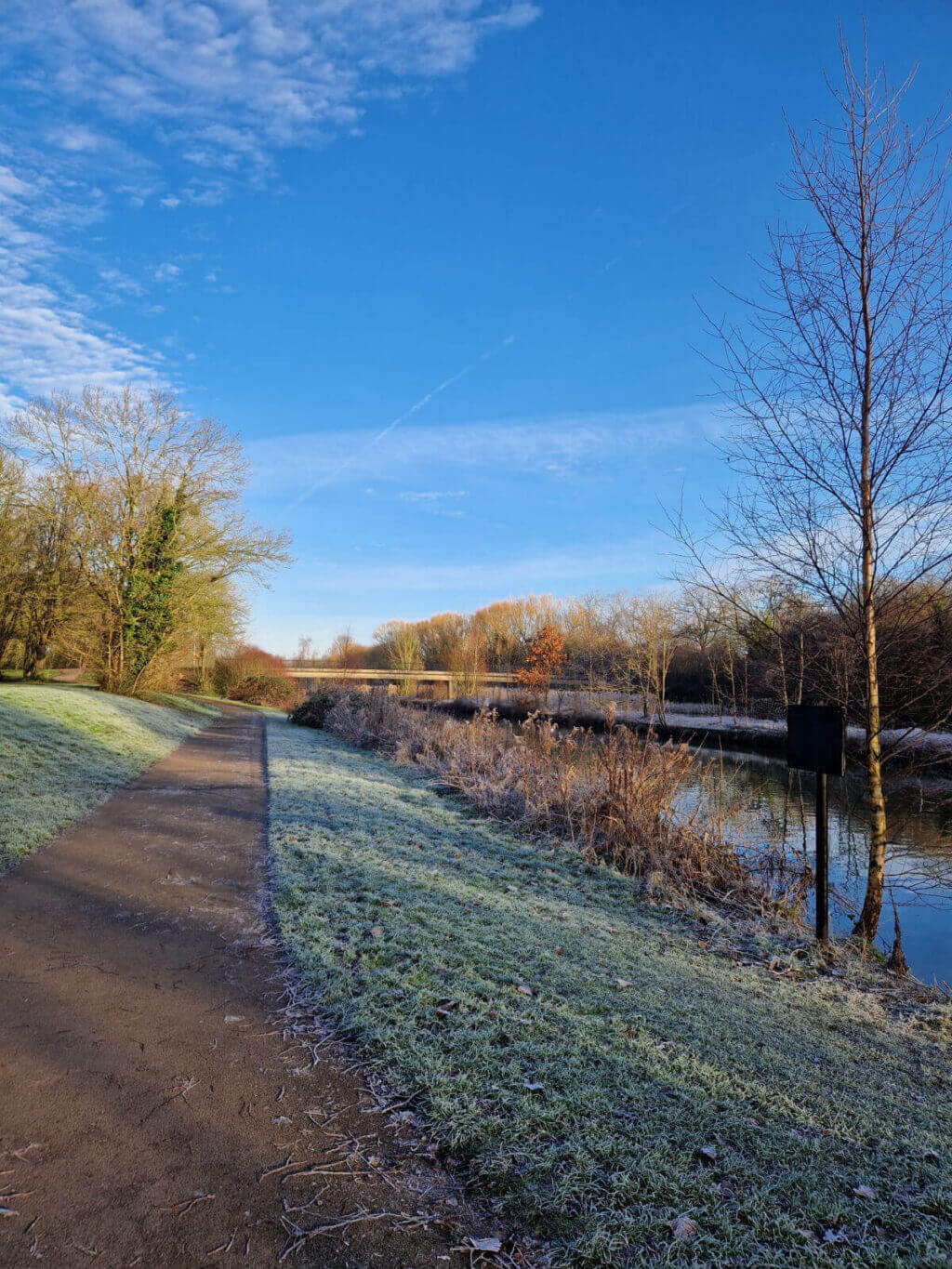 A gravel path near a canal on a frosty morning.  The grass is white with frost and the sky is blue and mostly cloudless