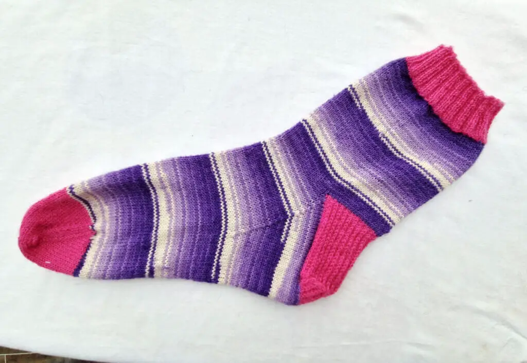 A single purple and white sock with pink cuffs, heels and toes lie on a white background. It has been pressed under a damp tea towel