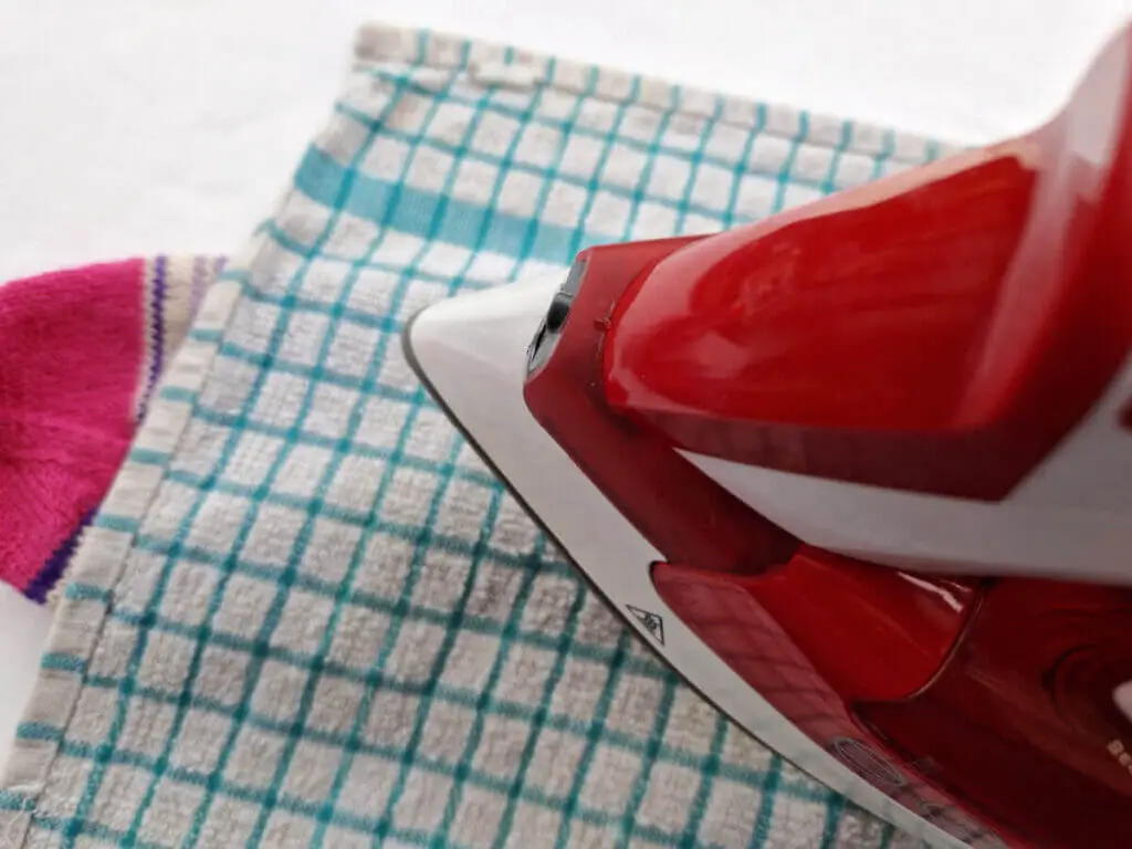 A red iron is over a wet tea towel covering a knitted sock
