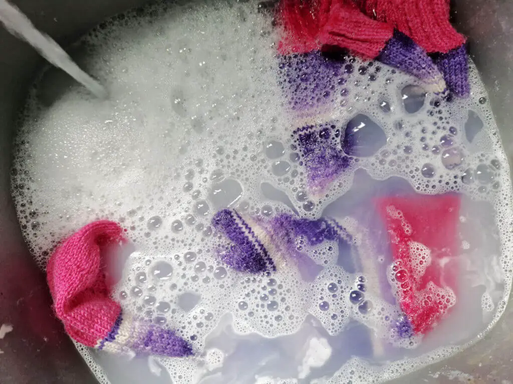 A pair of purple and white socks with pink cuffs, heels and toes soaking in a bowl of bubbly water