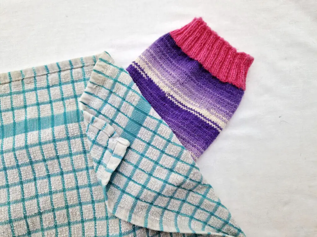 A single purple and white sock with pink cuffs, heels and toes lie on a white background. The sock is party covered by a blue and white checked tea towel