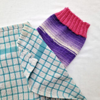A single purple and white sock with pink cuffs, heels and toes lie on a white background. The sock is party covered by a blue and white checked tea towel
