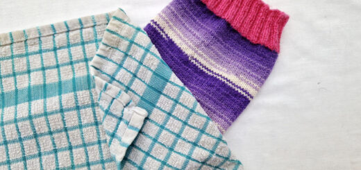 A single purple and white sock with pink cuffs, heels and toes lie on a white background. The sock is party covered by a blue and white checked tea towel