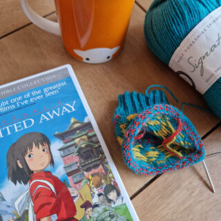 A partly knitted sock in turquoise, red and yellow lies next to a Spirited Away DVD case on a wooden table. There's an orange mug on the table too.