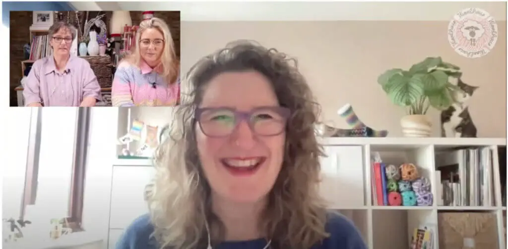 Christine is smiling into the camera on a video call.  In the top left corner is a shot of two ladies wearing shades of purple.  Behind Christine is a storage unit containing books, yarn and a tabby and white cat is hiding behind a plant