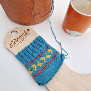 A partly-knitted blue sock on a wooden sock blocker, next to a leather project bag and an orange mug of tea. The socks have a band of golden fish in a red border around the top