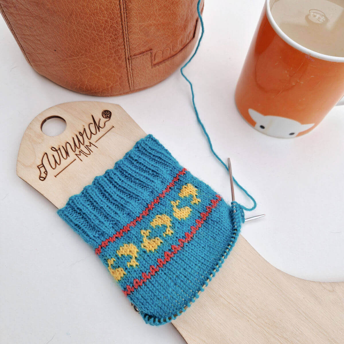 A partly-knitted blue sock on a wooden sock blocker, next to a leather project bag and an orange mug of tea. The socks have a band of golden fish in a red border around the top