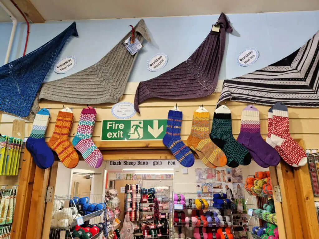 Happy Feet sock samples designed and knitted by Winwick Mum displayed over a doorway in Black Sheep Wools craft barn. Through the doorway are racks of yarn on sale, and above the socks are shawl samples