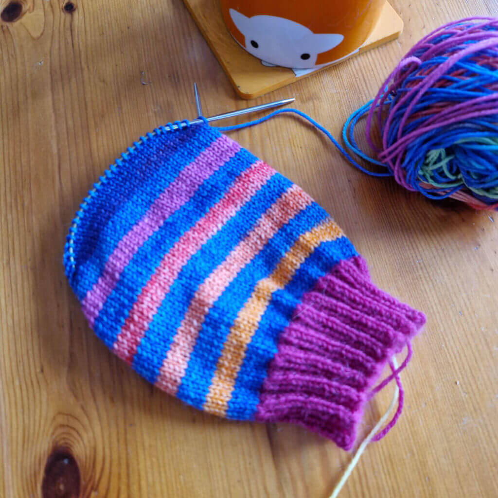 A partly knitted sock in rainbow shades broken up by bands of blue