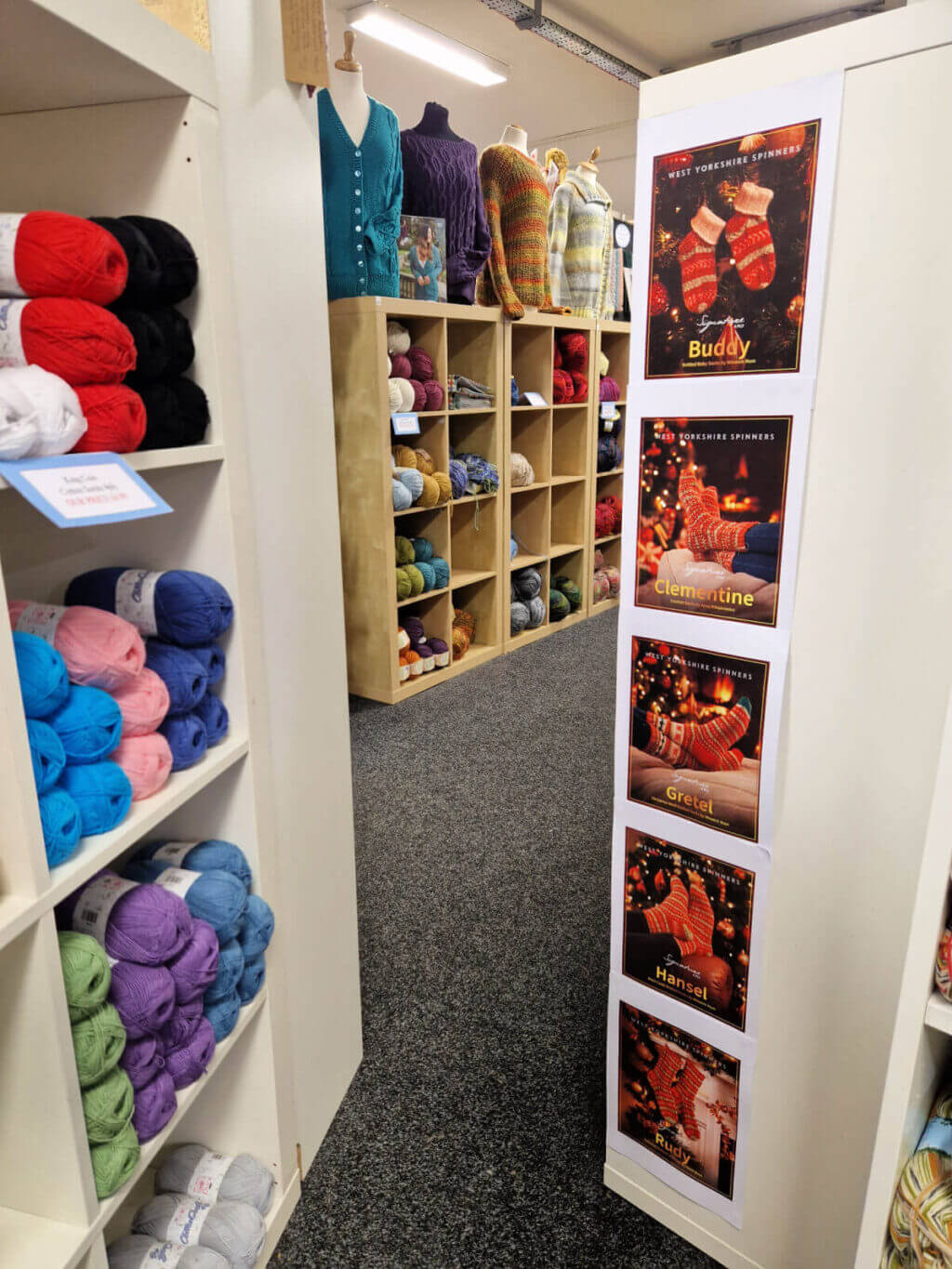 A view through shelving units to more units behind. Down the side of one of the units are photos of WYS Christmas socks patterns designed by Winwick Mum