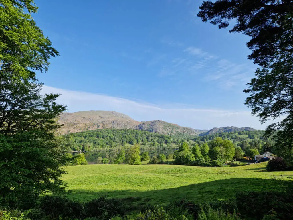 A closer view across the grass and lake to the mountain behind. The sky is blue and the water is very still, reflecting the mountain and trees