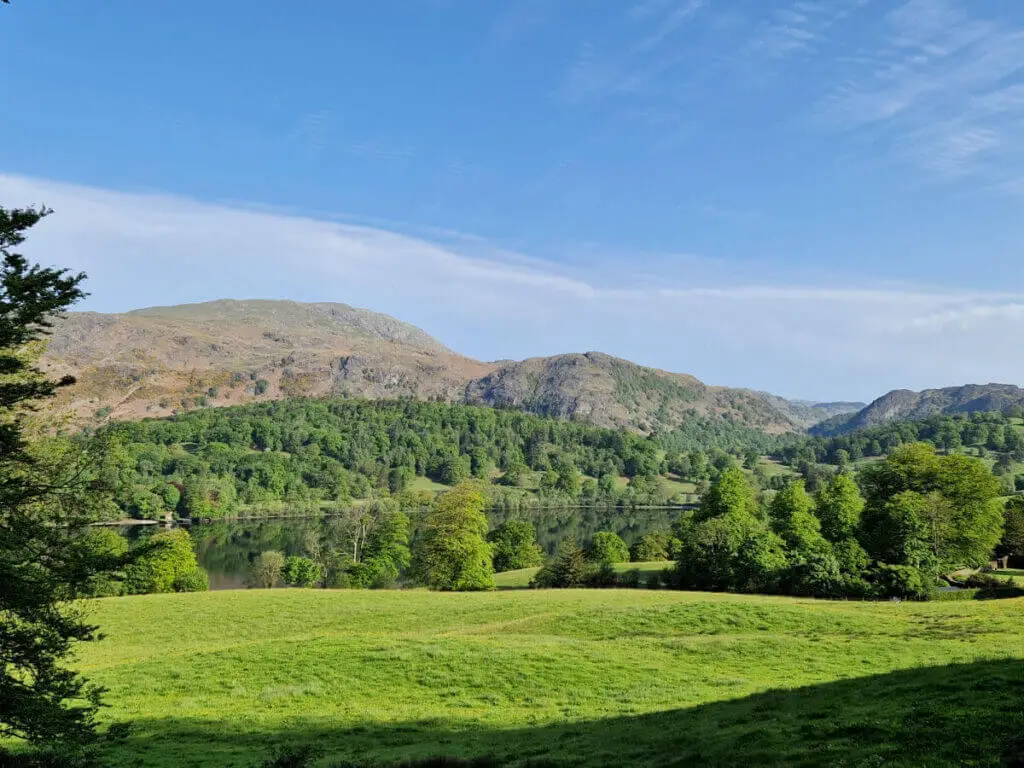 A view across the grass and lake to the mountain behind. The sky is blue and the water is very still, reflecting the mountain and trees