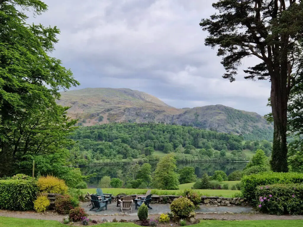 A more distant view across the grass and lake to the mountain behind. The sky is grey and the water is very still, reflecting the mountain and trees