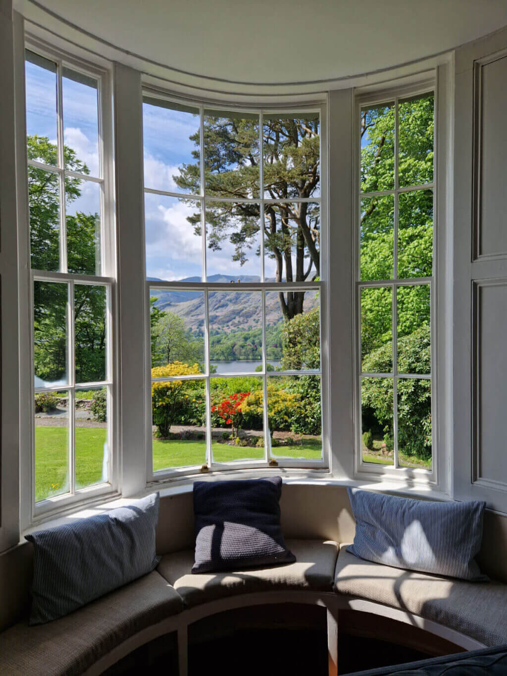 A view out of a bow window onto gardens and the mountains beyond. The sun is streaming through the window onto the cushions of the seat built into the window space