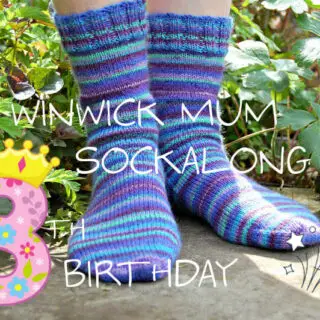 A pair of hand knitted blue striped socks modelled on feet which are standing on a stone flag in a flower border. The text across the picture reads Winwick Mum Sockalong 8th birthday