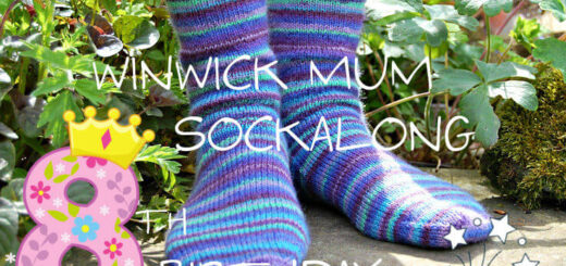 A pair of hand knitted blue striped socks modelled on feet which are standing on a stone flag in a flower border. The text across the picture reads Winwick Mum Sockalong 8th birthday