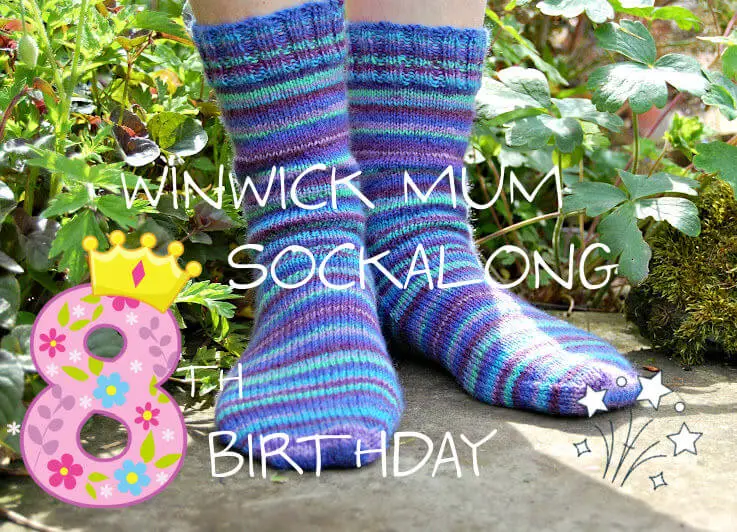 A pair of hand knitted blue striped socks modelled on feet which are standing on a stone flag in a flower border. The text across the picture reads Winwick Mum Sockalong 8th birthday