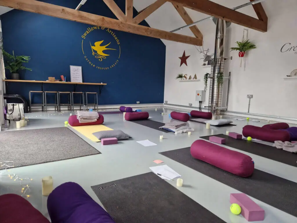 A view across a yoga studio laid out ready for a class. There are mats, bolsters, blankets and tennis ball accessories on the floor. The back wall is blue.
