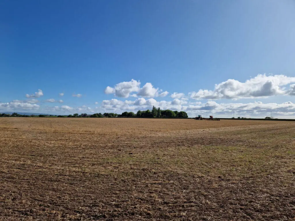 Looking across a brown field to trees in the distance. The sky is blue with white clouds and there is a tractor in the distance.