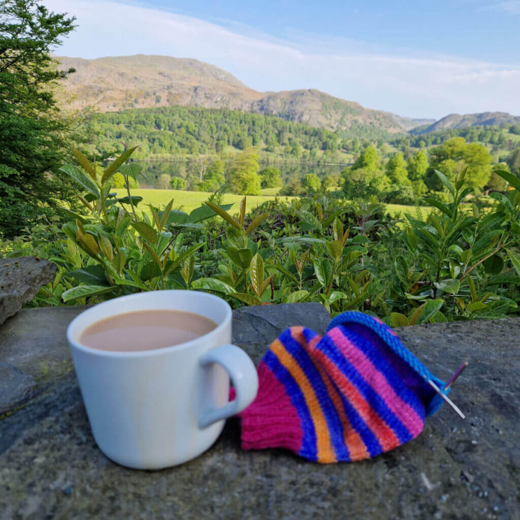 A white mug of tea and a rainbow-striped partly knitted sock are balanced on a stone wall. Behind them, the view is across green fields and trees to a lake and a mountain. The water is still and reflects the sky and the mountain