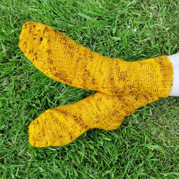 A pair of golden yellow socks with a twisted stitch pattern modelled on feet. The model is sitting on the grass, feet crossed at the ankle.
