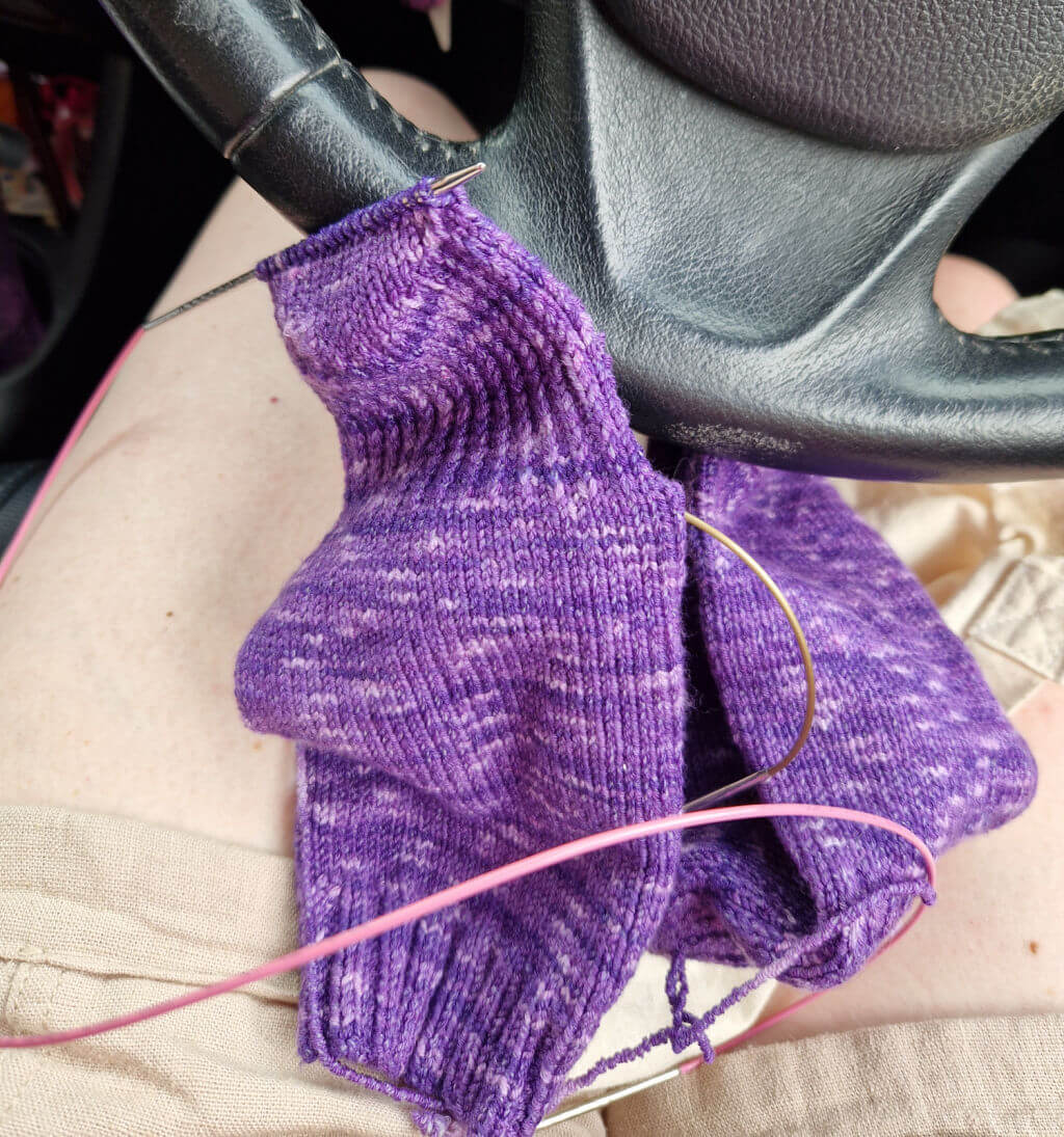 A partly-knitted purple sock rests on knees against a car steering wheel