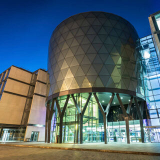 A futuristic-looking building with a round glass and silver-coloured tower between square ends is lit up against the night sky